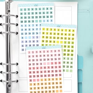 May include: A selection of colourful planner stickers, each with numbers 1-31. The stickers are in shades of blue, yellow, and pink. The stickers are arranged in a planner.