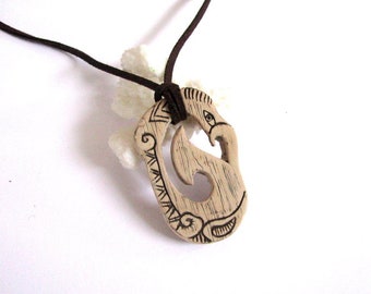 May include: A brown leather cord necklace with a carved wooden pendant. The pendant is shaped like a fish hook with intricate designs and a stylized bird head.