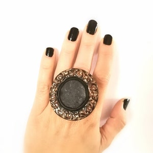 Puede incluir: Un anillo grande, redondo y negro con un diseño de filigrana dorada. El anillo se lleva en una mano con esmalte de uñas negro.