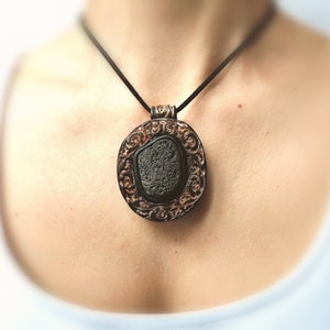 Puede incluir: Un collar con un colgante de piedra de lava negra y un marco de filigrana plateado. El collar está en un cordón negro.