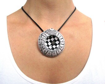 May include: A unique pendant necklace featuring a circular, silver-toned pendant with a black and white checkered center. The pendant has intricate detailing and hangs from a black cord. The necklace is worn against a white top.