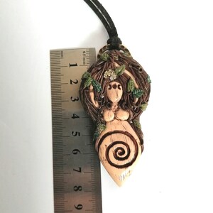May include: A brown and beige clay pendant necklace featuring a female figure with a spiral design and green leaves. The pendant is approximately 6 cm tall.