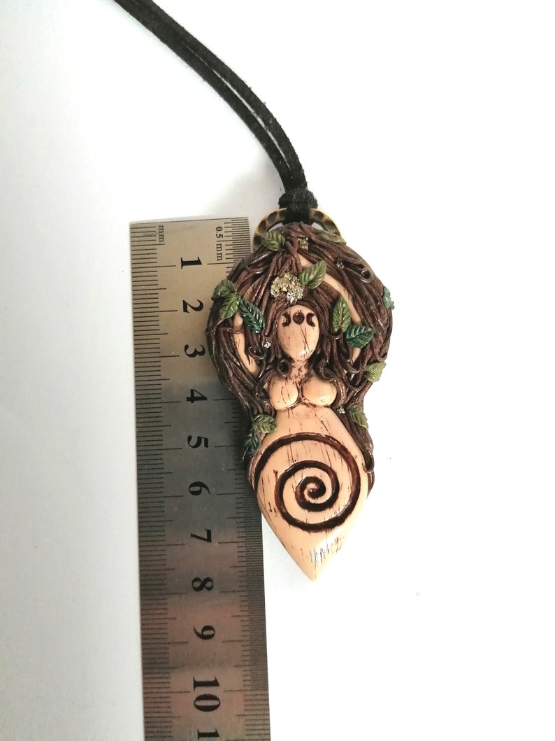 May include: A brown and beige clay pendant necklace featuring a female figure with a spiral design and green leaves. The pendant is approximately 6 cm tall.