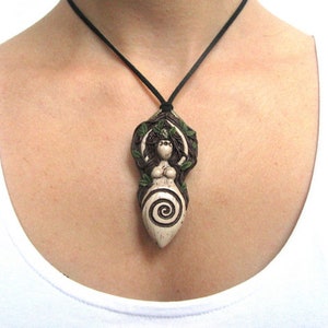 May include: A white ceramic pendant necklace featuring a depiction of a woman with long hair and a spiral design. The woman is surrounded by green leaves and vines. The necklace is on a black cord.