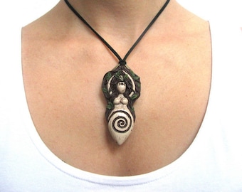 May include: A white ceramic pendant necklace with a depiction of a woman with long hair and a spiral design. The woman is surrounded by green leaves and vines. The necklace is on a black cord.
