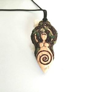 May include: A ceramic pendant necklace featuring a nude female figure with a spiral design on her belly, surrounded by brown branches and green leaves.