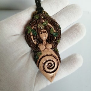 May include: A brown and green clay pendant depicting a woman with a spiral design and leaves. The pendant is on a black cord and has a gold-coloured metal bail.