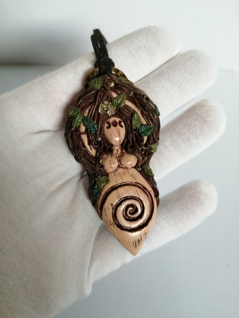 May include: A brown and green clay pendant depicting a woman with a spiral design and leaves. The pendant is on a black cord and has a gold-coloured metal bail.