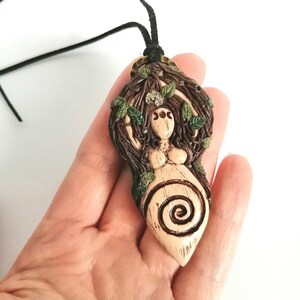 May include: A brown and green ceramic pendant depicting a woman with long brown hair, surrounded by leaves and branches. The pendant features a spiral design and is attached to a black cord.