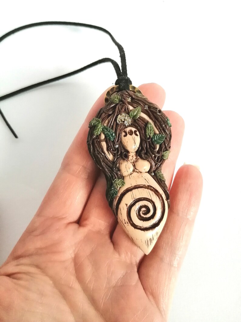May include: A brown and green ceramic pendant depicting a woman with long brown hair, surrounded by leaves and branches. The pendant features a spiral design and is attached to a black cord.