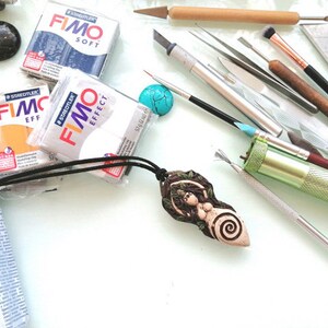 May include: A close-up of a polymer clay pendant with a nature-inspired design. The pendant is brown and green with a spiral pattern. The pendant is surrounded by various crafting tools, including sculpting tools, brushes, and a small turquoise stone.