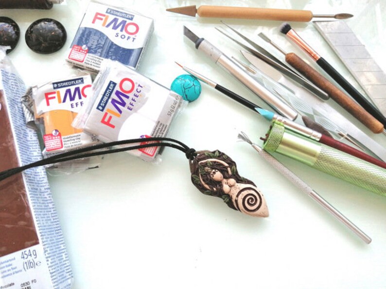 May include: A close-up of a polymer clay pendant with a nature-inspired design. The pendant is brown and green with a spiral pattern. The pendant is surrounded by various crafting tools, including sculpting tools, brushes, and a small turquoise stone.