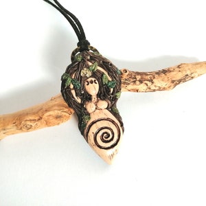 May include: A ceramic pendant necklace featuring a nude female figure with long brown hair, surrounded by green leaves and vines. The figure has a spiral design on her belly and three eyes on her forehead. The pendant is hanging from a black cord.