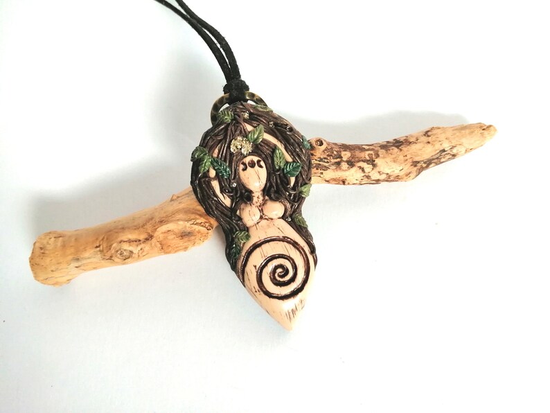 May include: A ceramic pendant necklace featuring a nude female figure with long brown hair, surrounded by green leaves and vines. The figure has a spiral design on her belly and three eyes on her forehead. The pendant is hanging from a black cord.