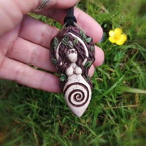 May include: A ceramic pendant featuring a woman with a spiral design, surrounded by leaves and vines. The pendant is brown, white, and green.