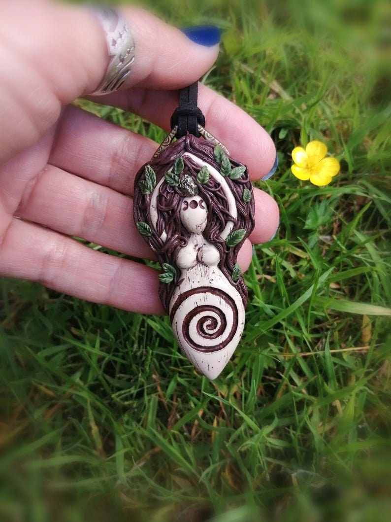 May include: A ceramic pendant featuring a woman with a spiral design, surrounded by leaves and vines. The pendant is brown, white, and green.