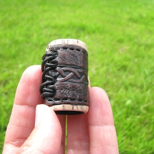 Op de afbeelding: Een bruin leren haarband met een gevlochten ontwerp en een witte rand. De haarband wordt in een hand gehouden.