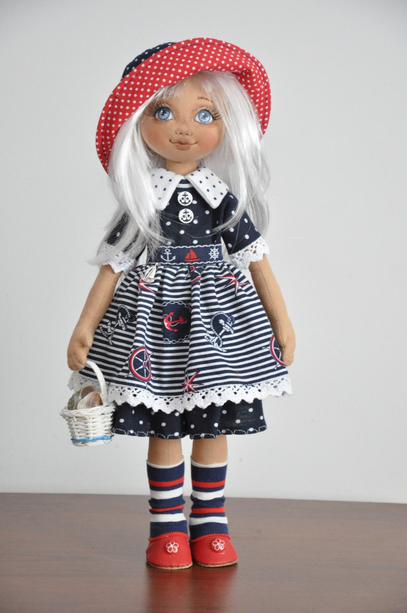 May include: A handmade doll wearing a blue and white striped dress with nautical themed prints, a red and white polka dot hat, and red and white striped socks. The doll is holding a white wicker basket.