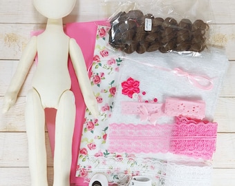 Op de afbeelding: Een poppenmaakset met een wit stoffen poppenlichaam, roze stof, witte stof met bloemenprint, bruin haar, roze en witte kantversiering, witte schoenen en roze bloemenversieringen.