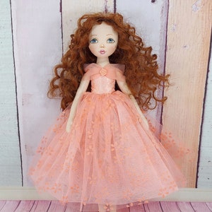 May include: A handmade doll with long, curly red hair wearing a peach-colored tulle dress with floral embroidery. The doll has blue eyes and a painted smile.
