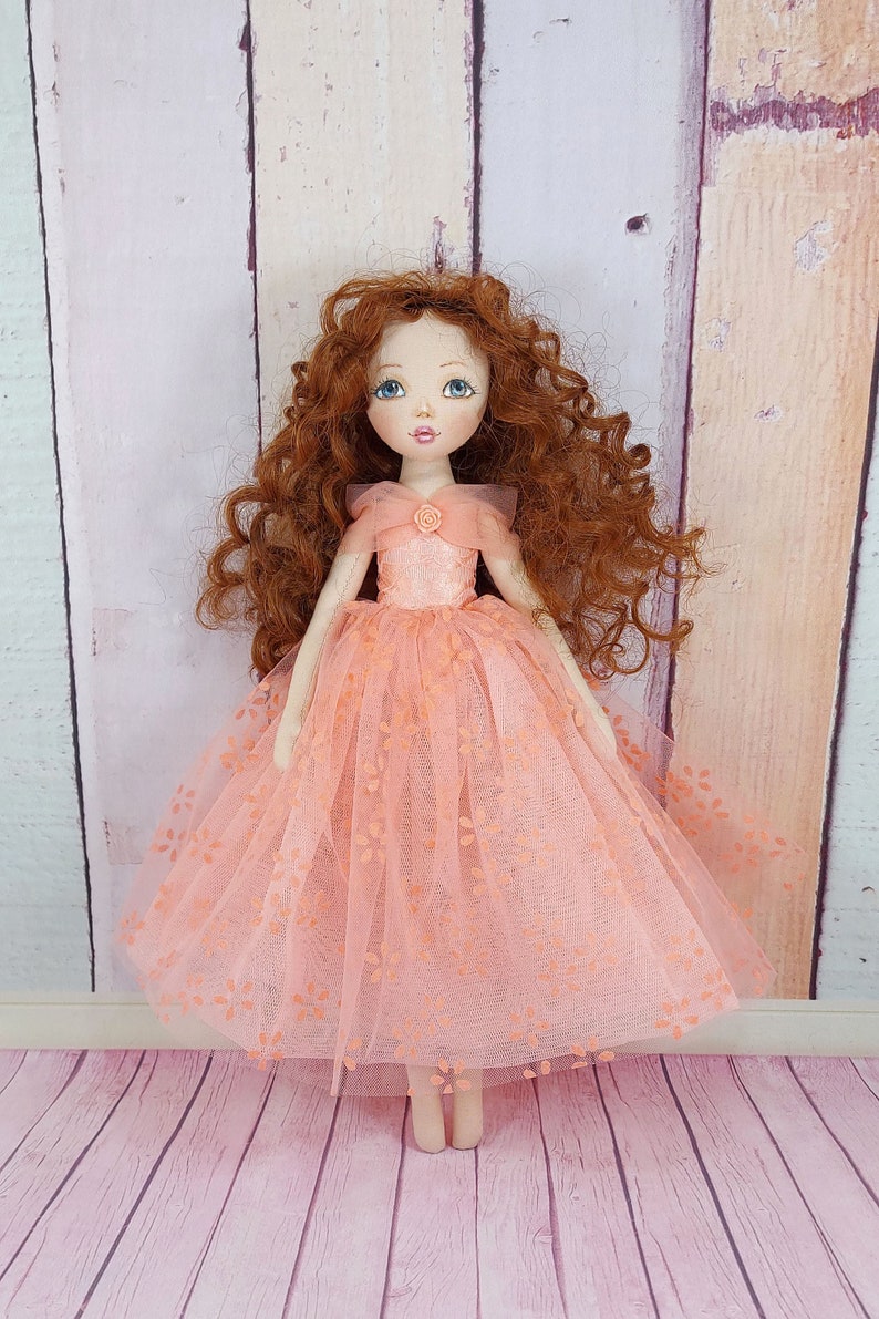 May include: A handmade doll with long, curly red hair wearing a peach-colored tulle dress with floral embroidery. The doll has blue eyes and a painted smile.