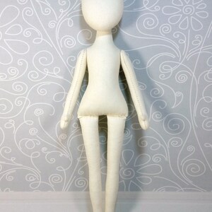 Op de afbeelding: Een witte stoffen pop met een eenvoudig ontwerp. De pop heeft een hoofd, armen, benen en een torso. De pop staat op een witte houten vloer.