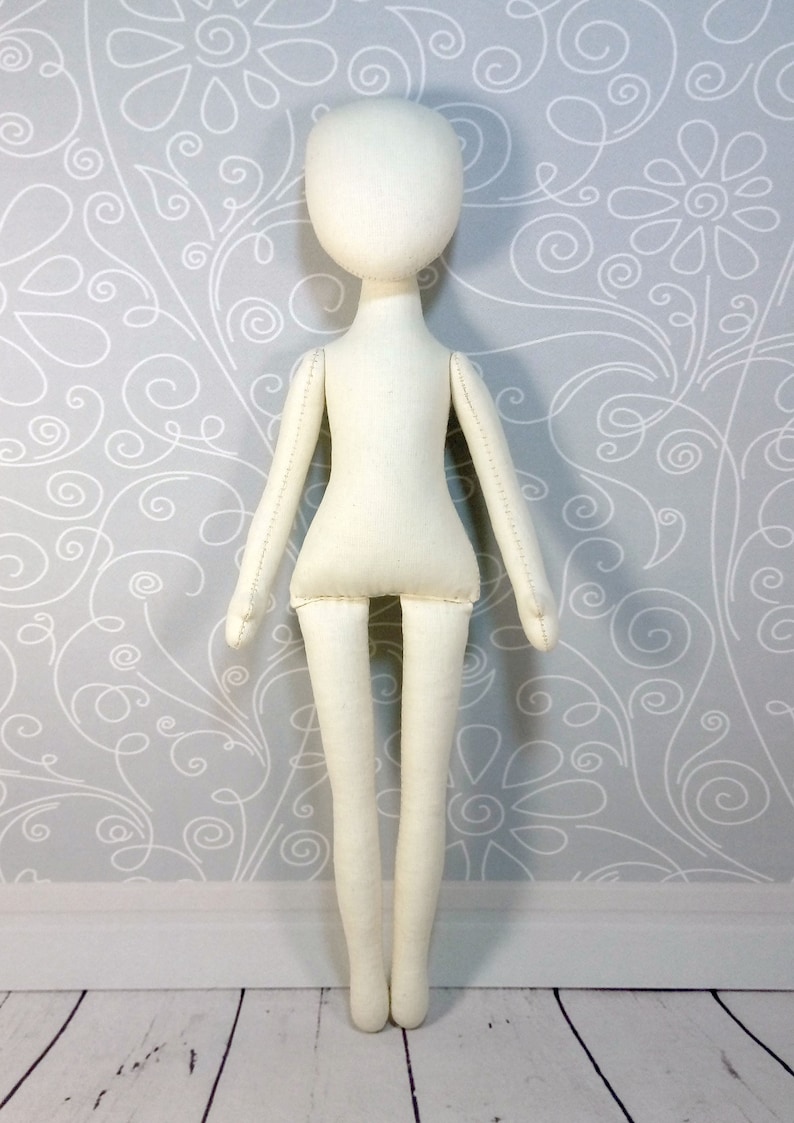 Op de afbeelding: Een witte stoffen pop met een eenvoudig ontwerp. De pop heeft een hoofd, armen, benen en een torso. De pop staat op een witte houten vloer.