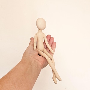 May include: A beige fabric doll with a round head and no facial features. The doll is sitting with its legs crossed and its arms outstretched.