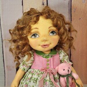Puede incluir: Una mu&ntilde;eca hecha a mano con cabello casta&ntilde;o rizado, vestida con un vestido rosa y verde y sosteniendo un oso de peluche rosa.