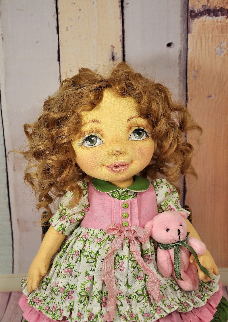 Puede incluir: Una mu&ntilde;eca hecha a mano con cabello casta&ntilde;o rizado, vestida con un vestido rosa y verde y sosteniendo un oso de peluche rosa.