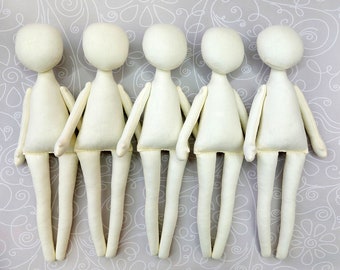 May include: Five unpainted white fabric doll bodies with long limbs. The dolls are arranged in a row on a patterned background.