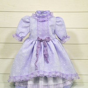 K&ouml;nnte beinhalten: Ein lavendelfarbenes Kleid mit wei&szlig;em Blumenmuster und Spitzenbesatz. Das Kleid hat einen R&uuml;schenkragen und eine Schleife an der Taille.