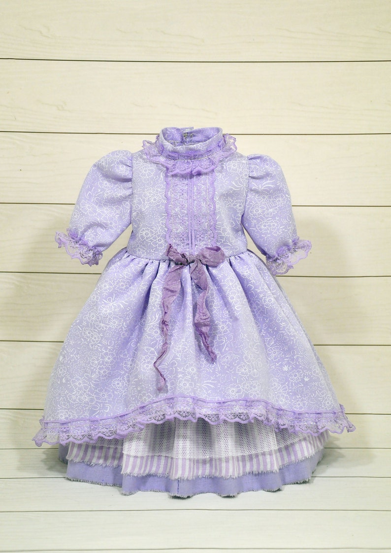K&ouml;nnte beinhalten: Ein lavendelfarbenes Kleid mit wei&szlig;em Blumenmuster und Spitzenbesatz. Das Kleid hat einen R&uuml;schenkragen und eine Schleife an der Taille.