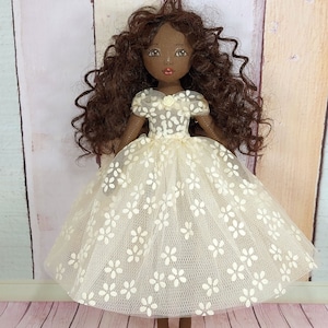 May include: A handmade doll with brown curly hair, wearing a white dress with floral embroidery. The doll has brown skin and painted features.