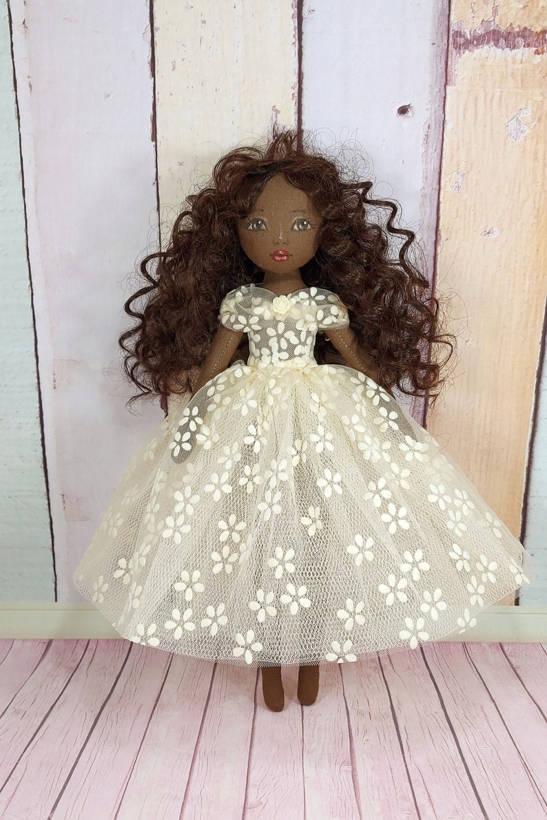 May include: A handmade doll with brown curly hair, wearing a white dress with floral embroidery. The doll has brown skin and painted features.