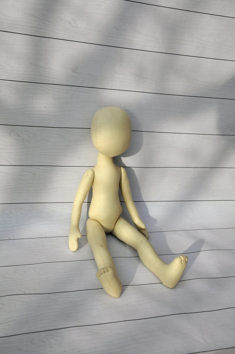 May include: A plain, unpainted, beige fabric doll with a round head and no facial features. The doll is sitting with its legs crossed and its arms resting on its legs.