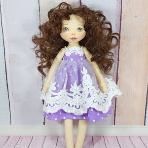 May include: A handmade doll with brown curly hair, wearing a purple dress with white polka dots and lace trim. The doll has blue eyes and a painted smile.