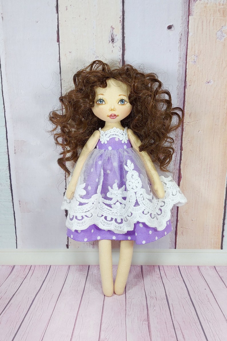 May include: A handmade doll with brown curly hair, wearing a purple dress with white polka dots and lace trim. The doll has blue eyes and a painted smile.