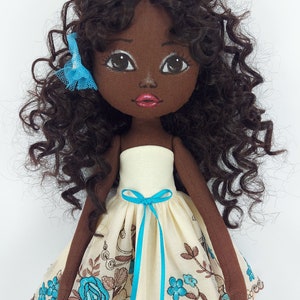 May include: A handmade doll with brown skin, curly brown hair, and a blue bow. She is wearing a white dress with blue floral embroidery and a blue ribbon.