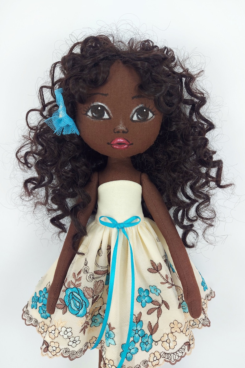 May include: A handmade doll with brown skin, curly brown hair, and a blue bow. She is wearing a white dress with blue floral embroidery and a blue ribbon.