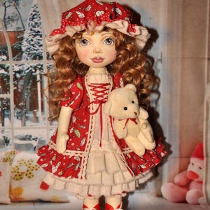 May include: A handmade doll with red curly hair, wearing a red and white dress with a white lace trim, a red and white hat, and red shoes. The doll is holding a white teddy bear.