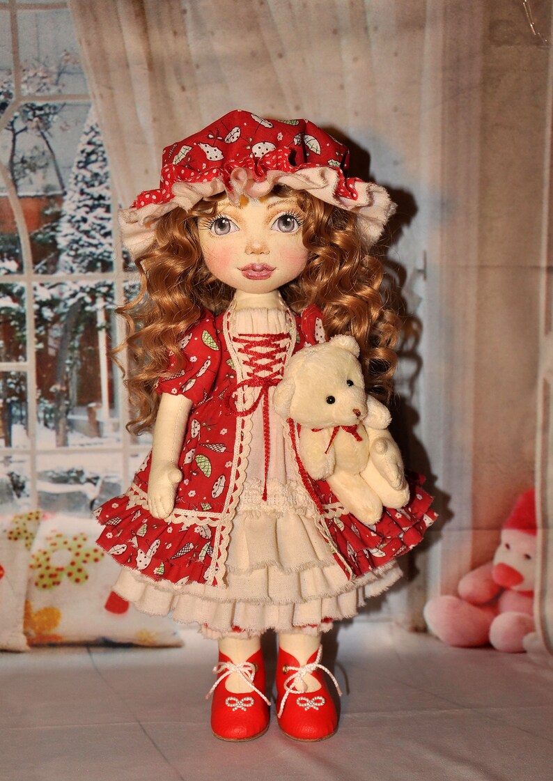 May include: A handmade doll with red curly hair, wearing a red and white dress with a white lace trim, a red and white hat, and red shoes. The doll is holding a white teddy bear.