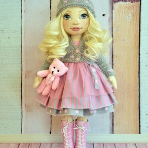 May include: A handmade doll with blonde hair, wearing a gray knitted hat, a pink and gray striped dress, a gray cardigan, and pink boots. The doll is holding a pink teddy bear.