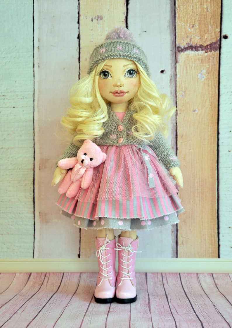 May include: A handmade doll with blonde hair, wearing a gray knitted hat, a pink and gray striped dress, a gray cardigan, and pink boots. The doll is holding a pink teddy bear.