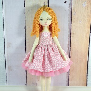 Op de afbeelding: Een handgemaakte pop met krullend blond haar, die een roze jurk met een hartjespatroon en een tule rok draagt. De pop draagt een kroon en heeft een lieve uitdrukking op haar gezicht.