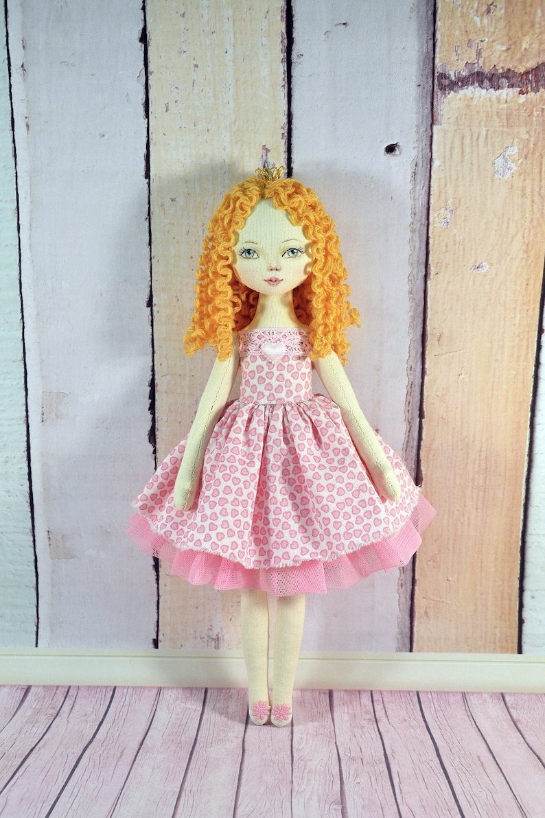 Op de afbeelding: Een handgemaakte pop met krullend blond haar, die een roze jurk met een hartjespatroon en een tule rok draagt. De pop draagt een kroon en heeft een lieve uitdrukking op haar gezicht.