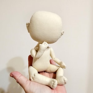 May include: A beige fabric doll with a large head and small limbs, sitting with its legs crossed. The doll is unpainted and has no facial features.