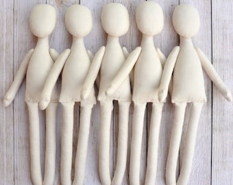 May include: Five unpainted white fabric dolls lined up in a row. The dolls are made of cotton fabric and have simple features. They are all the same size and shape.
