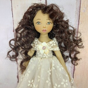 May include: A handmade doll with brown curly hair, wearing a white dress with a floral pattern and a white flower with a pink center on the bodice.
