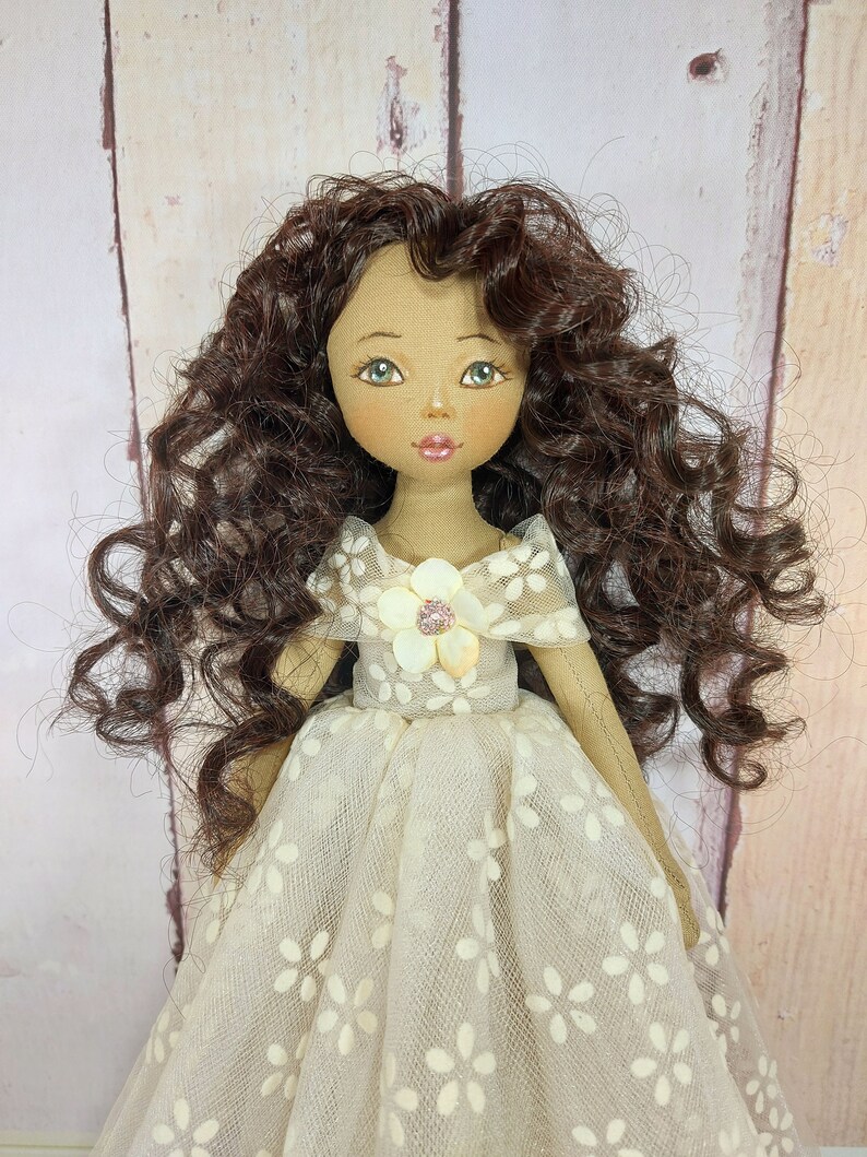 May include: A handmade doll with brown curly hair, wearing a white dress with a floral pattern and a white flower with a pink center on the bodice.
